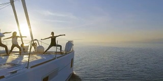 航拍侧视图拍摄的三个美丽的健康妇女做早晨瑜伽在一艘白色帆船双体船。平静的大海与升起的太阳和平静的大海。