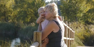 一位中年老奶奶抱着金发碧眼的小女孩站在自然公园的木桥上