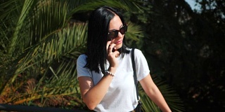 忙碌的女商人戴着墨镜在异国情调的棕榈树附近打电话。美丽的年轻女游客使用智能手机，与女友说话和微笑。缓慢的运动。多莉拍摄