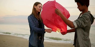 年轻的多民族夫妇举着红纸灯笼，准备出发。海滩上的浪漫约会。迷人的女人和她的非洲男朋友一起举着灯笼与火之前让它飞
