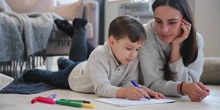 年轻漂亮的妈妈穿着暖和的毛衣躺在地板上，我的儿子用马克笔在纸上画他的家人。这孩子学画画。特写镜头