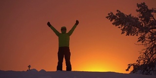 一个徒步旅行者站在白雪皑皑的山顶上，在日落的天空中自豪地伸出双臂