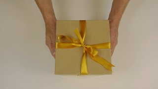 手上伸着一个礼盒，用金色的丝带系着一个蝴蝶结。送礼物。手捧礼盒祝贺某人的节日。赠送礼物。纸板箱是灰棕色的。视频素材模板下载