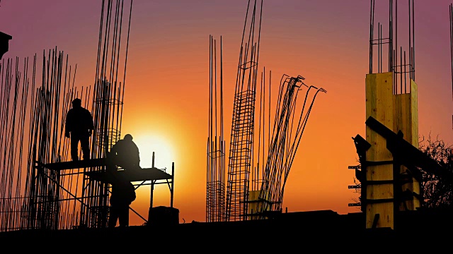 日落时分，在一处建筑工地，工人们在金属框架后面用铁丝将杆子固定在钢筋混凝土支柱的底部