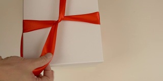 包装白色礼品盒。白种人手工包装礼盒。男人的手在一个白色的硬纸盒上系上一条红丝带。俯视图特写。红缎带缎带。
