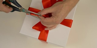 动作要快得多。包装白色礼品盒。白种人手工包装礼盒。男人的手在一个白色的硬纸盒上系上一条红丝带。俯视图特写。红缎带缎带。