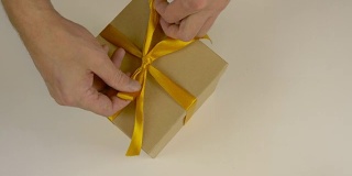 快速运动时间推移。包装礼盒。白种人手工包装礼盒。男人的手在一个棕色的硬纸盒上系上一条丝带。俯视图特写。金色的丝带，黄色的带子。