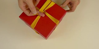 快速运动时间推移。包装红色礼品盒。白种人手工包装礼盒。男人的手在一个红色的硬纸盒上系上一条丝带。俯视图特写。金色的丝带，黄色的带子。