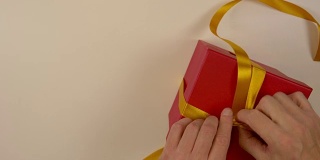 快速运动时间推移。包装红色礼品盒。白种人手工包装礼盒。男人的手在一个红色的硬纸盒上系上一条丝带。俯视图特写。金色的丝带，黄色的带子。