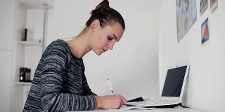 美丽的年轻女子坐在桌子旁，用她的钢笔写一封信。打开桌上的笔记本电脑。女商人在笔记本上做笔记，填写表格或文件