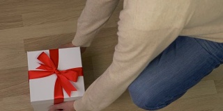 男子带来白色礼盒与红色丝带蝴蝶结，并把它放在地板上。木地板在室内。俯视图高角度。穿牛仔裤的男人拿着礼品盒走了。