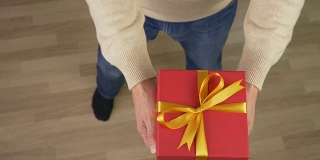 一名身穿米色毛衣的白人男子手里拿着一个礼品盒。俯视图的男人在休闲米色毛衣。人类给了礼物。锁定。红色的礼盒。高角度。铜。