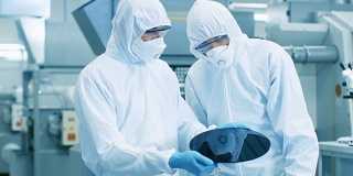 两名身穿无菌服的工程师/科学家/技术员手持将被制造成计算机芯片的半导体硅片。他们在一家现代半导体制造工厂工作。