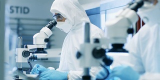 两名工程师/科学家/技术员穿着无菌洁净服使用显微镜进行组件调整和研究。他们在一家电子元件制造厂工作。