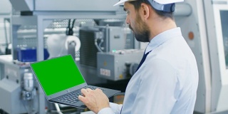 戴安全帽的工业工程师站在现代电子制造工厂的中央，在绿色模拟屏幕笔记本电脑上工作。