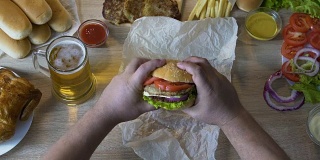 胖男人的手打开新鲜的汉堡，把它送到他的嘴，油腻的食物