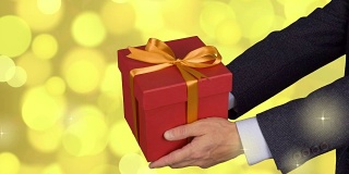 两只男性的手拿着红色的礼盒，上面有金色的蝴蝶结。庆祝除夕礼物礼盒。穿着古典西装的白人。金灯的背景。锁定。