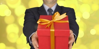 一个穿西装打领带的男人拿着一个红色礼品盒。男人给予礼物。一个手里拿着礼物的商人。背景抽象金色闪光闪烁，圈光散焦。