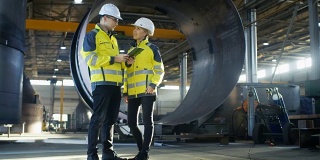 男和女工业工程师在安全帽讨论新项目，同时使用平板电脑。他们会做一些有表现力的手势。他们在重工业制造厂工作。长针。