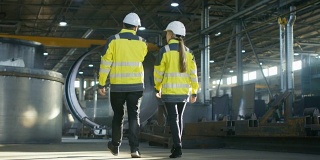男工程师和女工程师在重工业制造厂边走边讨论的背影照片。大型金属制品结构，管道元素躺在周围。