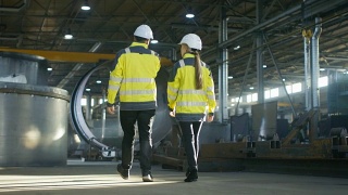 男工程师和女工程师在重工业制造厂边走边讨论的背影照片。大型金属制品结构，管道元素躺在周围。视频素材模板下载