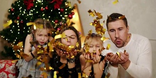 年轻幸福的四口之家坐在圣诞树旁，吹着金色的五彩纸屑。可爱的妈妈，爸爸和两个女儿一起庆祝圣诞节。Slowmotion拍摄