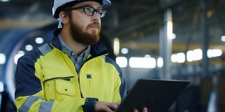 工业工程师戴安全帽穿安全外套使用笔记本电脑。他在重工业制造工厂工作，各种金属加工工艺正在进行中。