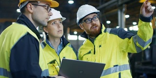 三个工业工程师在使用笔记本电脑时与工厂工人交谈。他们在重工业制造工厂工作。