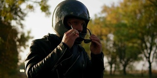 一个年轻人穿着黑色皮夹克和头盔戴着太阳镜，而坐在他的摩托车上准备开始他的旅程在森林。慢动作镜头