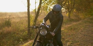 一个戴着头盔、穿着皮夹克的年轻人走到他的自行车前，在一个阳光明媚的秋天站在路边时启动了引擎。Slowmotion拍摄