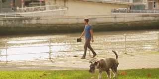 一个人在河边散步，他的狗在草坪上跑，河上的桥