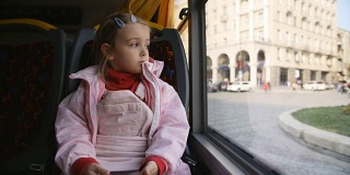 穿着粉红色夹克的金发小女孩，孩子坐在巴士车厢里。坐在她的座位上，透过玻璃窗看。
