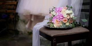 美丽的新娘花束在桌子上与美丽的婚纱和新娘饰品的背景。婚礼礼服在肩膀上，仪式前。美丽的婚礼花束和新娘的礼服