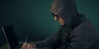 程序员戴着帽子和眼镜在笔记本电脑键盘上打字。黑客写的备忘录。男人用手做手势。低调的侧视图黑客坐在桌子前的笔记本电脑。