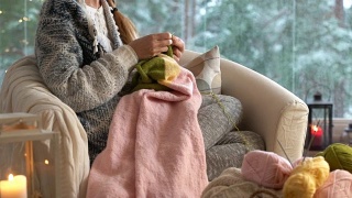 年轻女子在客厅里织着温暖的羊毛衫，窗外是雪景视频素材模板下载