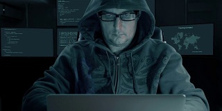 戴着时尚眼镜戴着兜帽的白人男子为互联网编程。来自笔记本显示器屏幕的Man tale stiker。房间里有许多后台显示器