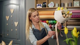 女金发花商安排现代花束和完成它与完美的缎带。英俊的花店老板在她的花店制作花束视频素材模板下载