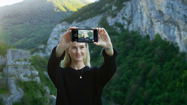 美丽的金发女孩用她的智能手机自拍，拿着屏幕对着相机。在背景风景优美的青山和山景。
