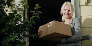 一个拿着包裹的妇女站在她家的门槛上。看着镜头，微笑着。包裹送货上门