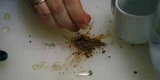 这名男子断开了凝胶状的药片，将其中的药片洒在了桌子上。一种干燥的褐色物质从其中沉淀出来。然后，他把药溶解在一杯水里。
