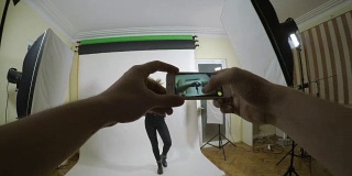 一个年轻的摄影师用智能手机在一个白色背景的工作室拍摄她的模特女友的观点