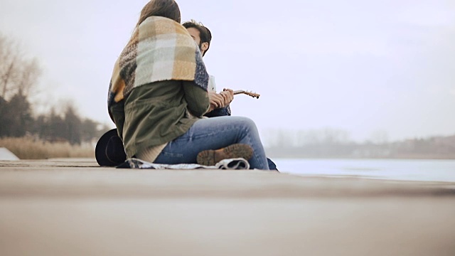 一个年轻人坐在结冰的湖边的码头上给他的女朋友弹奏尤克里里琴