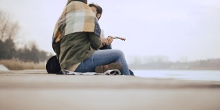 一个年轻人坐在结冰的湖边的码头上给他的女朋友弹奏尤克里里琴