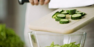 特写女性的手切片新鲜黄瓜，并把它加入沙拉碗