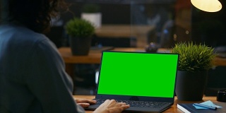 美丽的办公室女员工在她的桌面工作在一个模拟绿屏笔记本电脑。过肩镜头。晚上创意办公室。