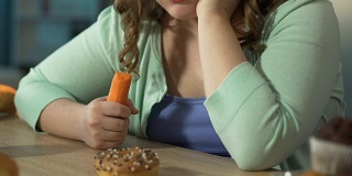 体重超标的女孩一边吃新鲜胡萝卜，一边悲伤地看着桌上的甜甜圈