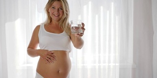 肖像孕妇饮用矿泉水在玻璃和摩擦裸露的腹部靠近窗户