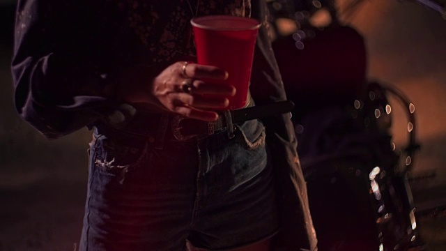 年轻的骑摩托车的女人拿着塑料杯在晚上的音乐节