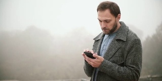 年轻英俊的男子站在雾蒙蒙的清晨公园的肖像。胡须男使用带触摸屏的智能手机