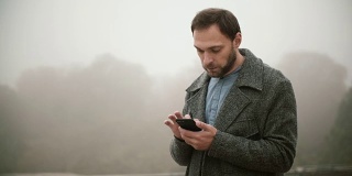 年轻英俊的男子在雾蒙蒙的清晨早早地站在公园里。胡须男在用带触摸屏的智能手机